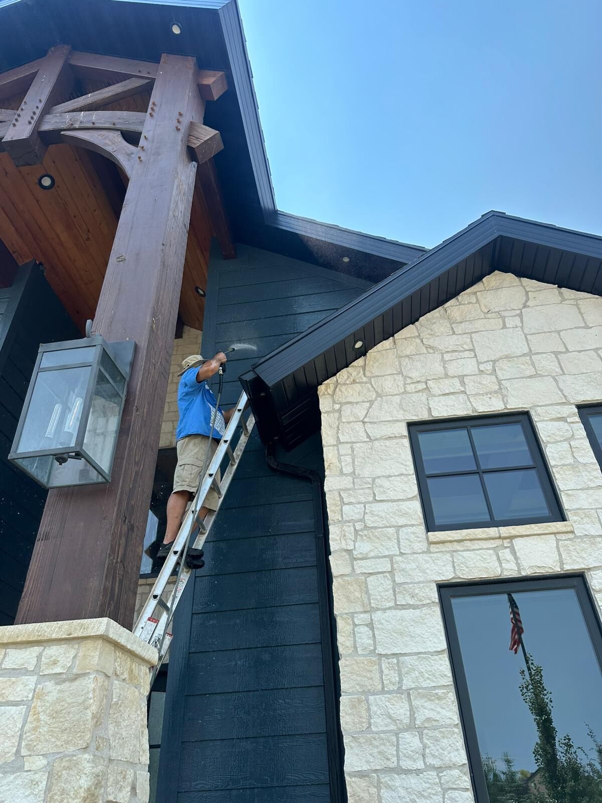 Nice Glass technician on a ladder soft washing dark lap siding under a timber-framed porch, next to a cream stone gable with black-framed windows