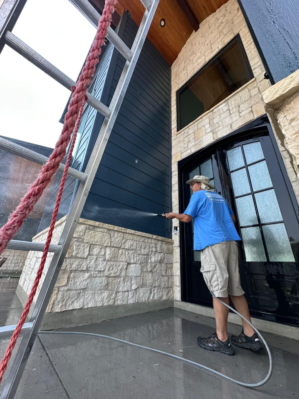 Nice Glass technician rinsing dark lap siding above a stone wainscot at a home's front entry, beside tall glass double doors