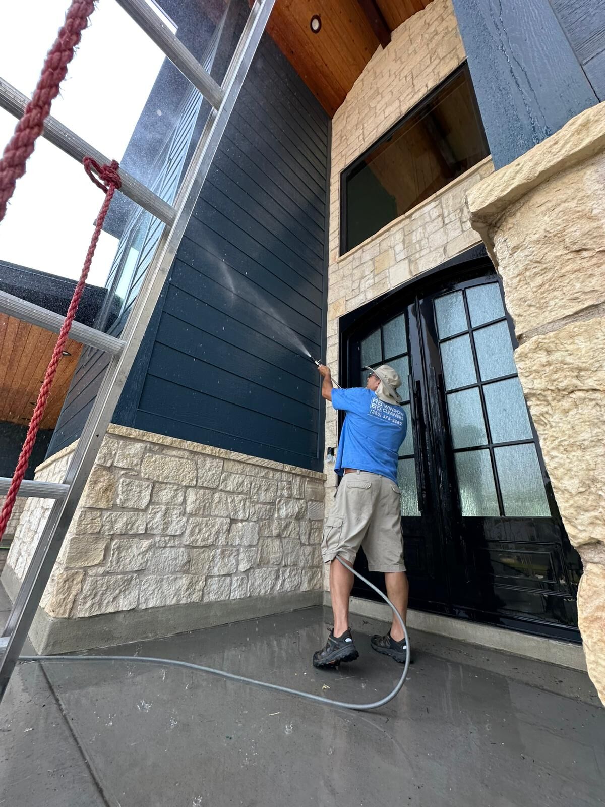 Nice Glass technician soft washing tall dark lap siding above a stone wainscot beside a home's black arched front doors